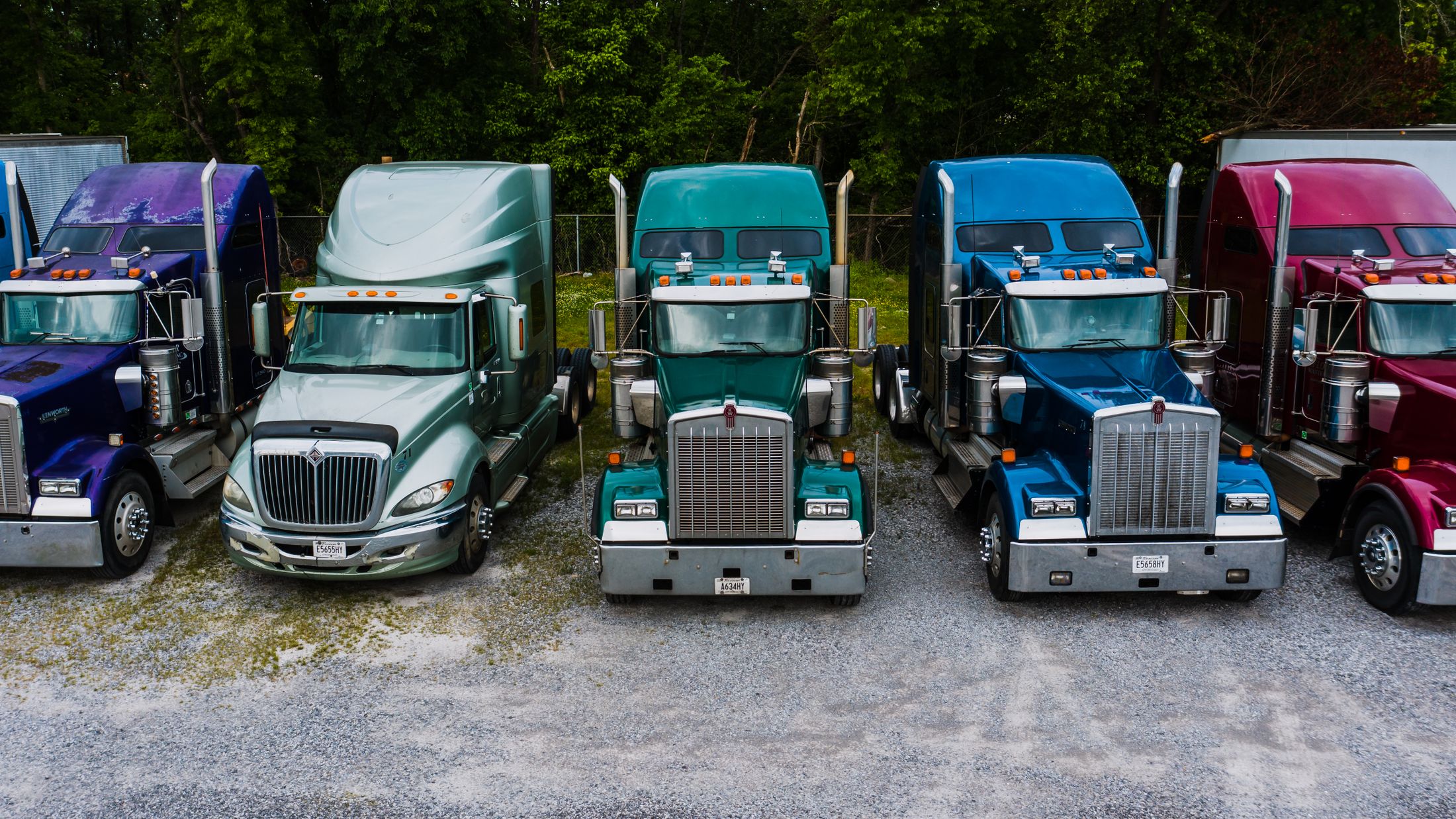 American semi-truck cabs parked in a row at a gravel depot backed by green trees.