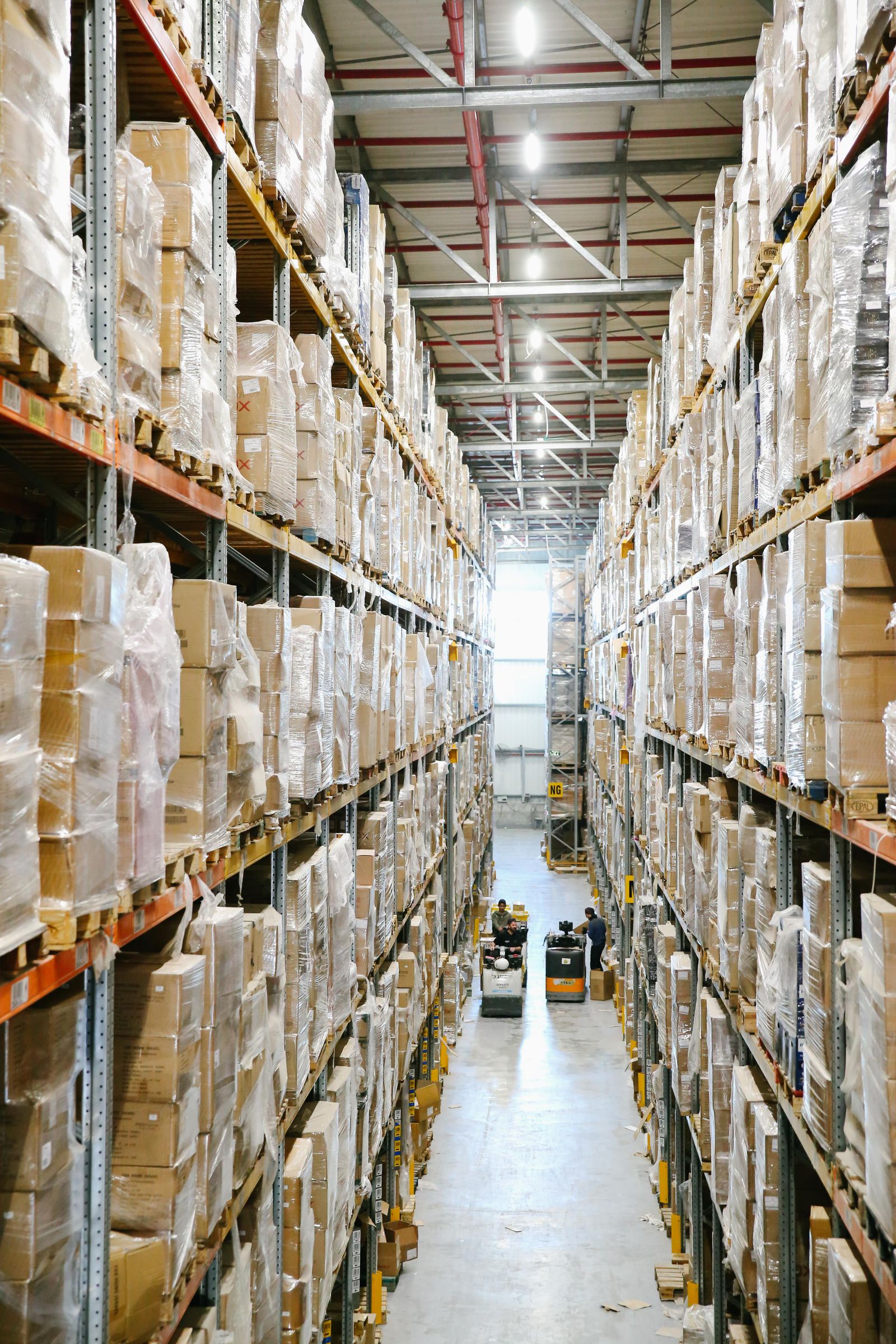 A tall warehouse aisle lined with shrink-wrapped cartons on pallet racks, with workers and pallet equipment in the distance.