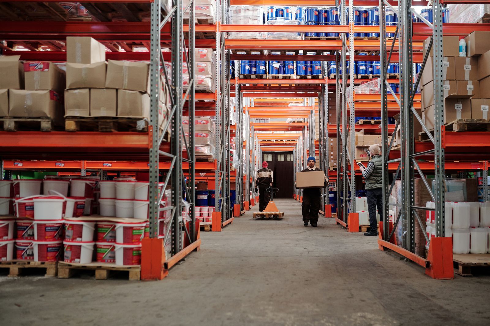 A warehouse worker carrying a cardboard carton while colleagues manage inventory and a pallet jack in the aisle.